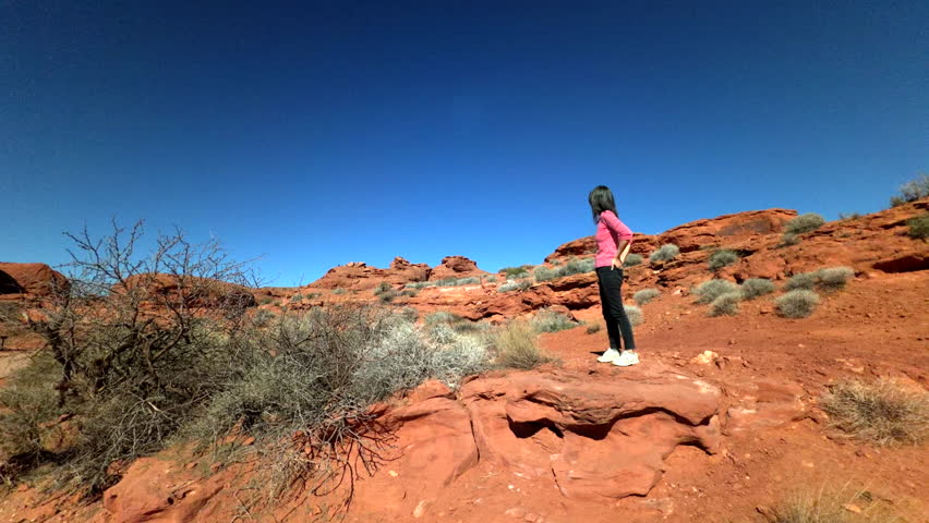 Looking up at woman standing on rugged terrain near vibrant red boulders of  sandstone formation deep blue sky background 60FPS 4k