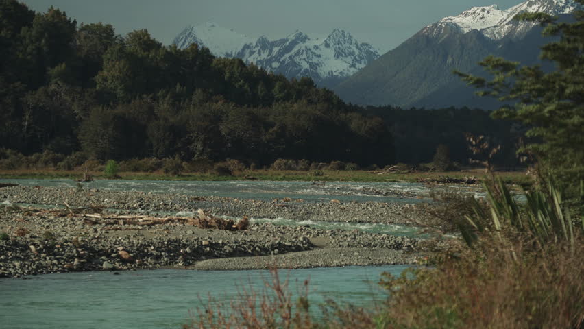 Icy blue water flows over rocks in its path under a New Zealand forest and mountain landscape.