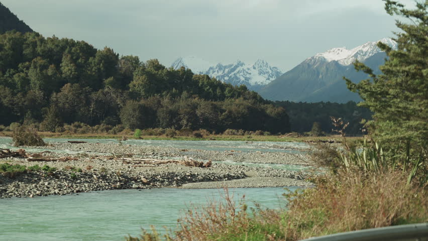 Water flows over a glacial river plain beneath a dark forest and snow capped mountains in New Zealand.