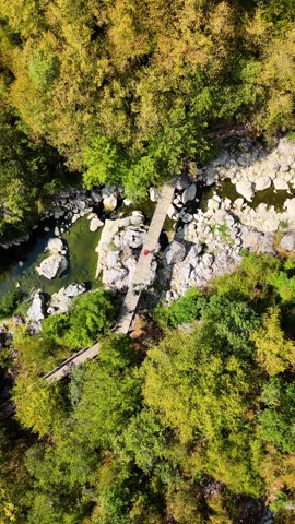 Cinematic top-down aerial drone view of Horma Canyon in Pınarbaşı Kastamonu Turkey, captured at a 90 degree angle looking straight down as the drone slowly descends over a suspension bridge with three unrecognizable people walking above a narrow river flowing through steep rocky cliffs and dense forest