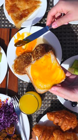 Top down view of a person spreading delicious orange jam on a slice of toast with a knife, enjoying a lavish breakfast buffet with croissants, juice, and fruits on a sunny morning