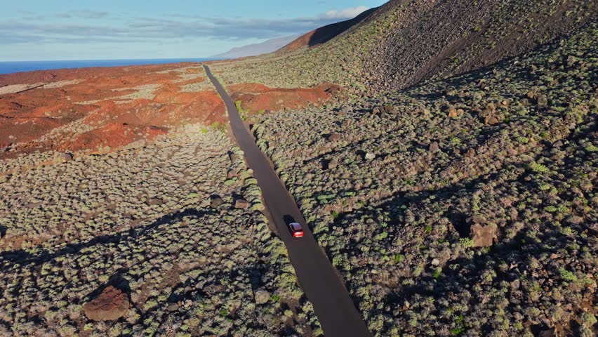 Car drives along lonely asphalt road through spectacular red volcanic fields of El Hierro. Rugged desert landscape at sunset, Canary Islands, Spain. Volcanic road trip on El Hierro island