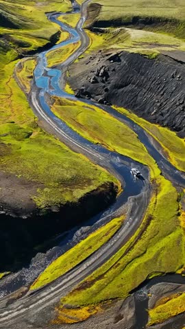 Vertical drone view of an S-shaped river beside a gravel road as a 4x4 crosses the Iceland highlands, surrounded by bright moss and dark volcanic slopes.