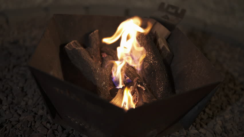 Firewood flames rising from wood logs in metal fire pit with intense heat and dark night atmosphere, medium orbit