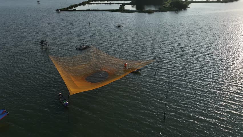 An aerial orbit shot captures a traditional Vietnamese fisherman operating a square lift net on the serene Thu Bon River during golden hour, revealing the rural landscape.