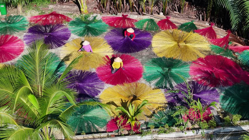 Aerial pull-out reveals vibrant sedge grass drying in circular patterns at a traditional Vietnamese craft village, with workers and lush palm trees by a river.