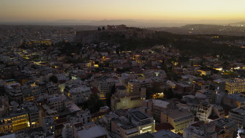 Dusk aerial over Athens with Acropolis hill emerging as city lights begin to glow softly, panoramic orbit