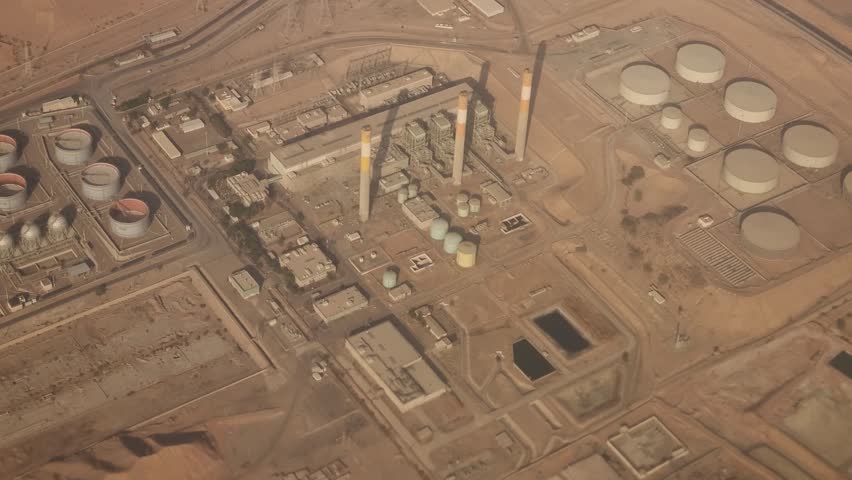 Aerial view from an airplane over Jordan showing a large industrial complex in the desert, likely an oil refinery or gas processing plant, with storage tanks, pipelines, and tall chimneys