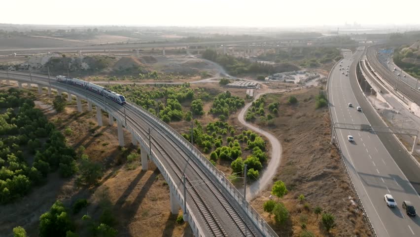 Aerial view of a modern passenger train crossing a high viaduct bridge over a busy highway in Israel. Cinematic 4K transportation and infrastructure footage.
