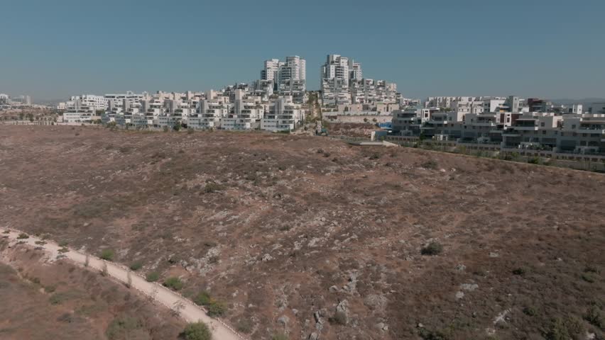 4K drone footage of a modern residential neighborhood in Israel built on a hillside. The video features white terraced apartment buildings, high-rise towers, and arid landscape,