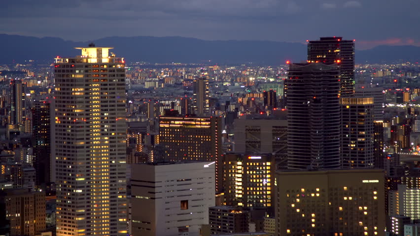 A zoomed-in aerial perspective of the Umeda business district and Nakanoshima skyline at sunset. Features the glowing windows of office towers and the dense urban grid of Osaka in winter