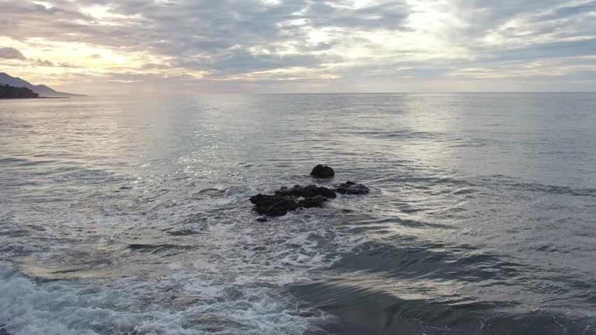 Stunning aerial view of a golden sunrise over the sea with soft waves hitting dark rocks, featuring a dramatic cloudy sky and misty horizon.
