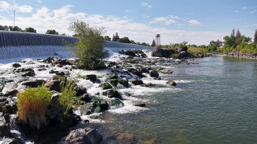 Aerial shot of the Snake River with water cascading over a rock barrier into a calmer pool, surrounded by vegetation and blue sky.