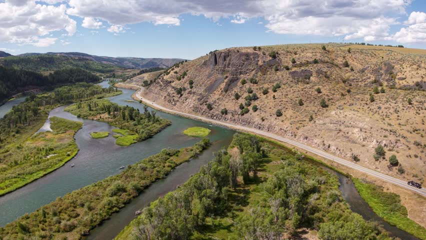 Aerial footage of the Snake River cutting through a rocky canyon in Idaho, with green vegetation along the riverbanks and rugged hillsides under a partly cloudy sky.