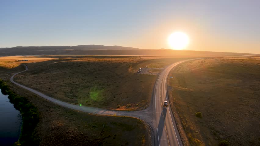 Aerial view of a winding road through Idaho farmland at sunrise, with the sun low on the horizon casting golden light across the fields and hills.