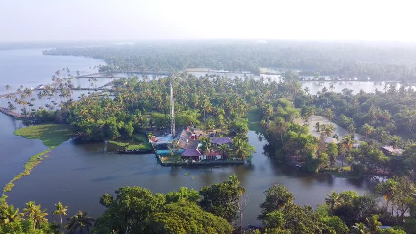 Aerial view of a serene waterfront temple and traditional villa nestled in the lush tropical backwaters of Kerala, surrounded by palm trees and tranquil lagoons in golden morning light.