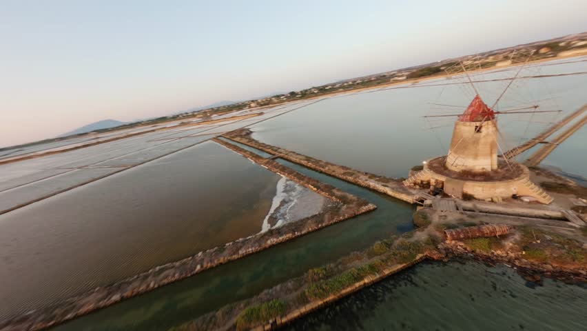 FPV drone zoom in over Salina salt flats at sunset, revealing a traditional windmill, salt ponds and warm golden light reflecting on water.