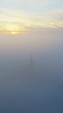 Aerial view of the Stevenskerk standing above Nijmegen during a hazy sunrise, with gentle mist moving through the narrow streets and golden light coloring the sky. The church tower emerges from the fo