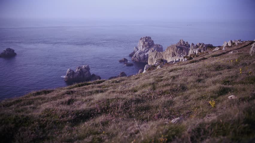 Finistere coastline in Brittany featuring rocky cliffs and calm ocean waters during blue hour. Ground level view of grassy slopes leading to the sea in France.