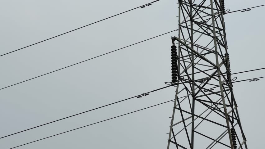 Close-up view of a porcelain insulator mounted on a steel lattice transmission tower, with parallel power lines crossing the frame against an overcast sky.
