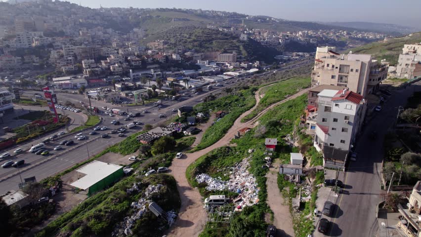 High aerial drone shot of an Arab town in the Wadi Ara region of Israel, showing dense hillside housing, main roadway traffic, surrounding green valleys, and mixed urban–rural landscape under daylight