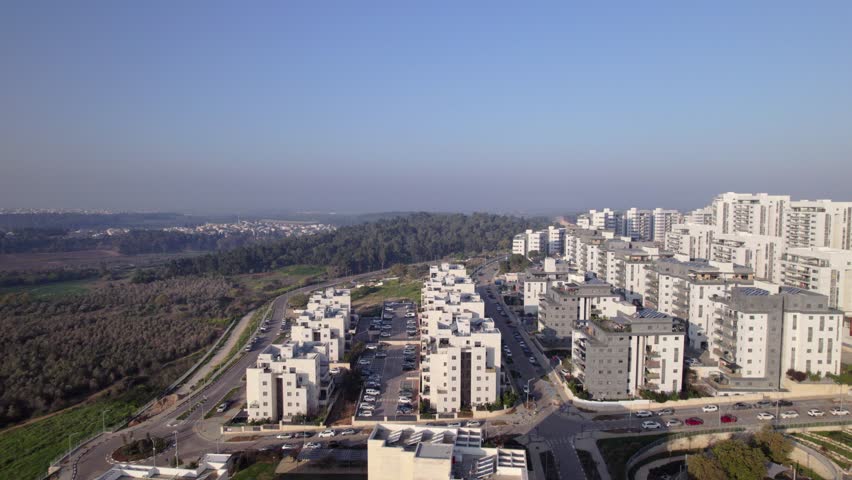 High aerial drone shot of the city of Harish in Israel, showing modern residential buildings alongside a natural oak forest and curving roadway under clear daylight.