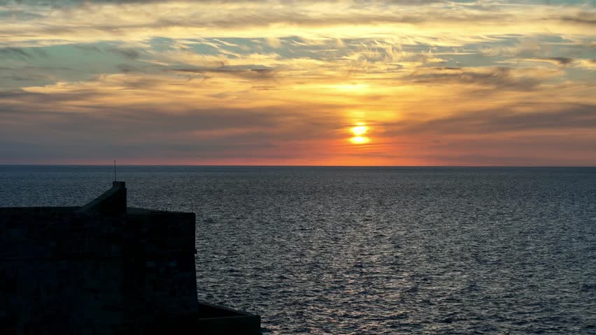 Breathtaking golden sunset over the Mediterranean Sea seen from the historic stone ramparts of Gozo Malta during a calm autumn evening with a wide angle view of orange glowing clouds deep blue water