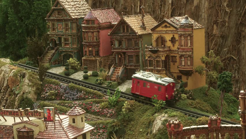 Vintage Miniature Toy Train Set With A Red Caboose Electric Engine Climbing A Mountain In A Country Town.