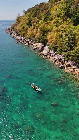 Traditional Thai longtail boat approaching rocky tropical shoreline with lush green jungle vegetation in Phuket, Thailand. Aerial drone view over crystal clear turquoise Andaman Sea, iconic island travel scene