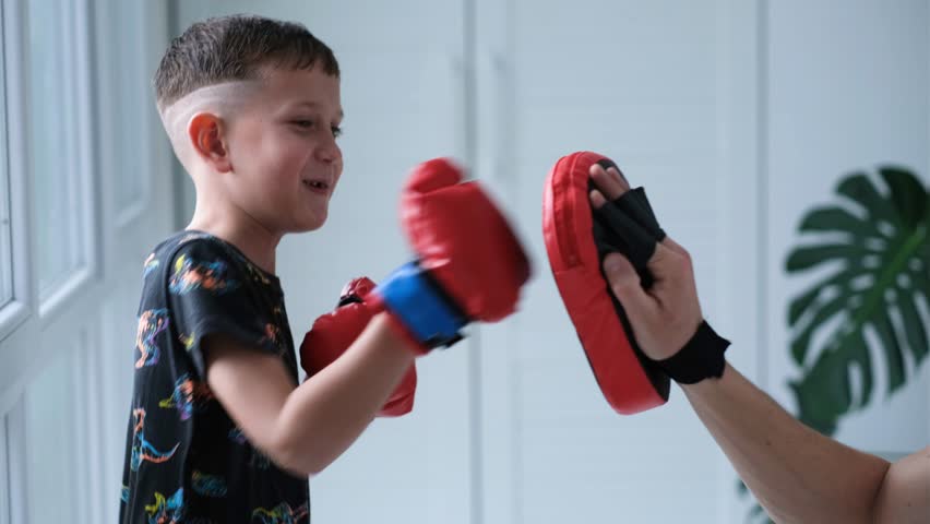 A child wearing boxing gloves practices boxing with his father. The boy punches the boxing mitt.
