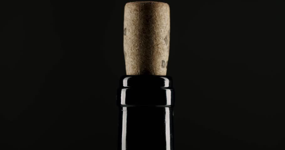Close-up of a hand carefully pushing a natural cork into the neck of a dark glass wine bottle against a black background, representing the process of bottling and sealing wine