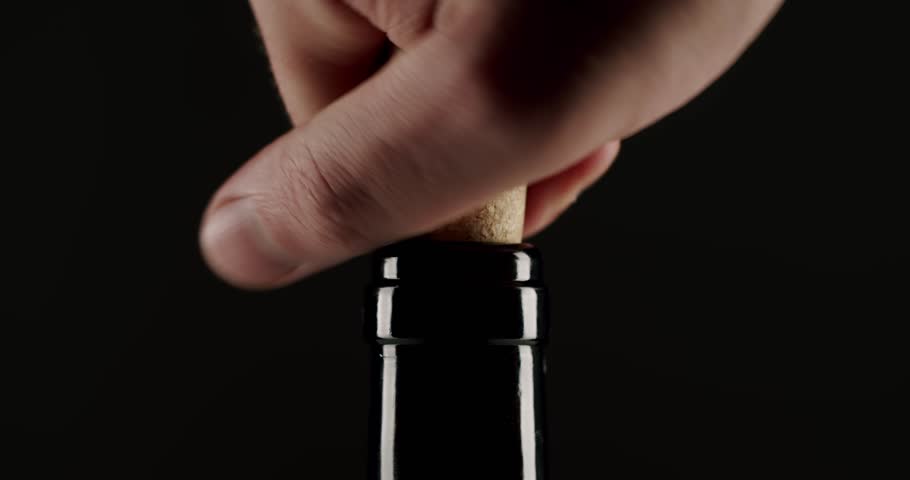 Close-up of a hand carefully pushing a natural cork into the neck of a dark glass wine bottle against a black background, representing the process of bottling and sealing wine