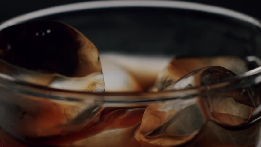 Close-up of black coffee pouring into a glass filled with ice cubes against a dark background, making a refreshing cold beverage perfect for a hot summer day in slow motion
