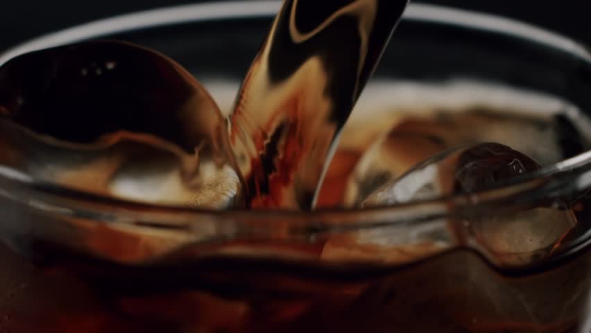 Close-up of black coffee pouring into a glass filled with ice cubes against a dark background, making a refreshing cold beverage perfect for a hot summer day in slow motion