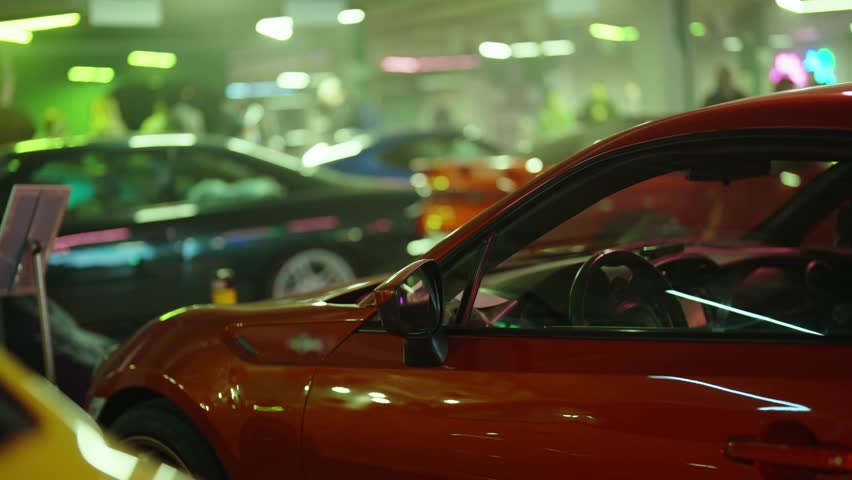 Modern red sports car shining under vibrant green and purple neon lights at a motor exhibition. With other customized vehicles and spectators blurred in the background creating an exciting atmosphere