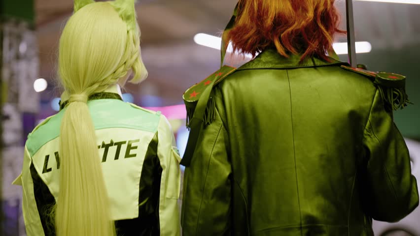 Two women in intricate anime character costumes walking from behind at a comic convention, one with blonde hair and cat ears and the other with red hair, a leather jacket and a sword
