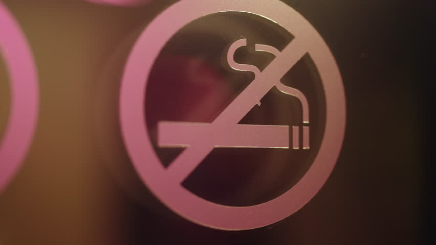 Intimate slow-motion shot of a no smoking sign sticker on a glass surface, with blurry figures and warm city lights moving and reflecting in the background at night, creating an atmospheric feel