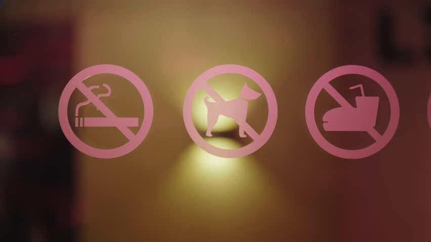 Pink round prohibition signs indicating no smoking, no dogs, and no food or drinks are stuck on a glass door, with a warm, blurry, out-of-focus background gently moving
