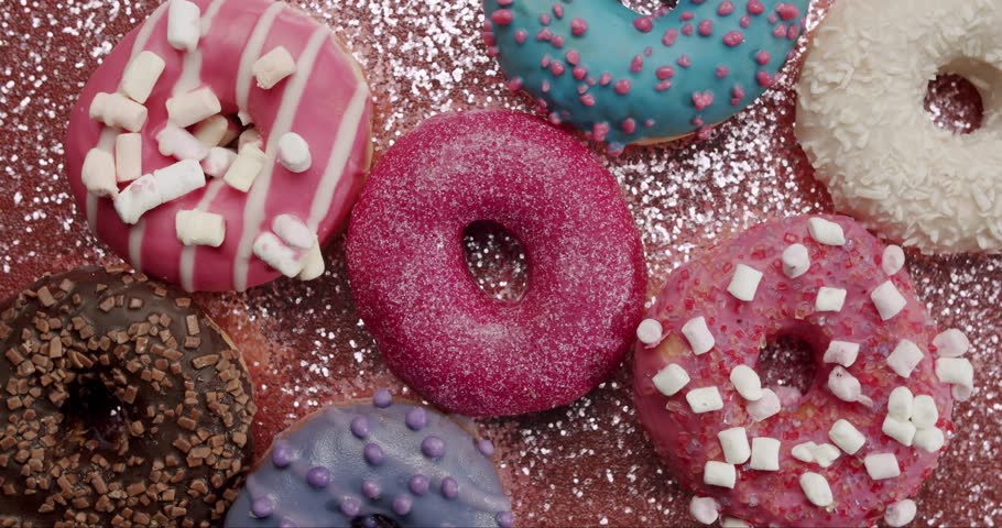 Delicious and colorful donuts with various toppings and icings on a sparkling pink background while powdered sugar gently falls, creating a sweet and festive culinary composition