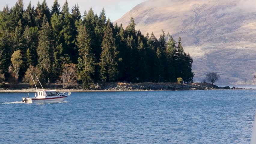 Landscape scenery of fishing charter boat travelling along Lake Wakatipu inlet with bushland forest and mountain headland in the distant background Otago Region New Zealand tourism nature and travel