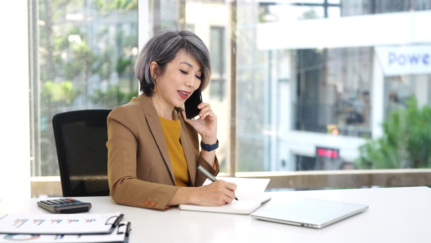 Mature middle aged business woman, 50s professional lady executive manager talking on the phone making business call on cellphone and taking note at work in office .