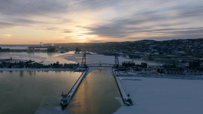 High-altitude panoramic aerial of the Duluth harbor and Lift Bridge during an arctic sunset. Sea smoke swirls over the frozen lake while city lights begin to glow.