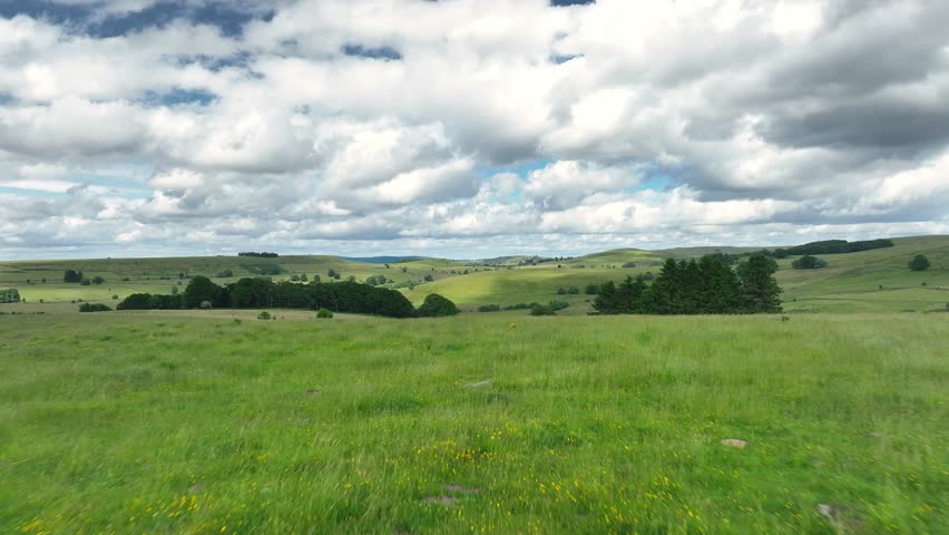 Scenic aerial drone footage of lush green pastures and rolling hills on the Aubrac plateau in France, capturing the peaceful rural countryside under a cloudy sky.