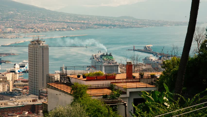 Scenic view of Naples harbor featuring large cruise ship at port and coastline. Coastal cityscape with buildings, sea view, and distant mountains seen from elevated terrace.