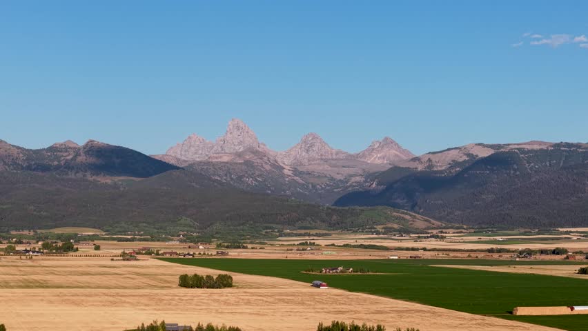Drone view of Idaho farmland with golden fields and green pastures framed by distant mountain peaks under a wide blue sky