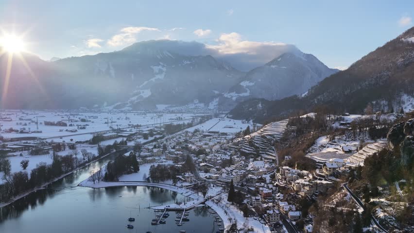 Walensee Switzerland drone flying over Walenstadt town, snowy Swiss alps and calm alpine lake under morning sun.