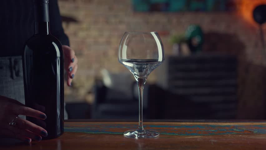 Elegant woman pouring red wine form wine bottle to a wine glass. Tasting and drinking wine at home in a dark cosy room, Focus in front on glass. 4K UHD video clip.