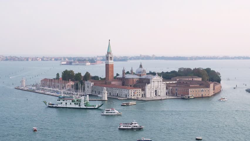 Elevated viewpoint of Venice, Italy, featuring the San Giorgio Maggiore church overlooking the lagoon, with historic architecture surrounded by ocean waters under a bright holiday atmosphere.