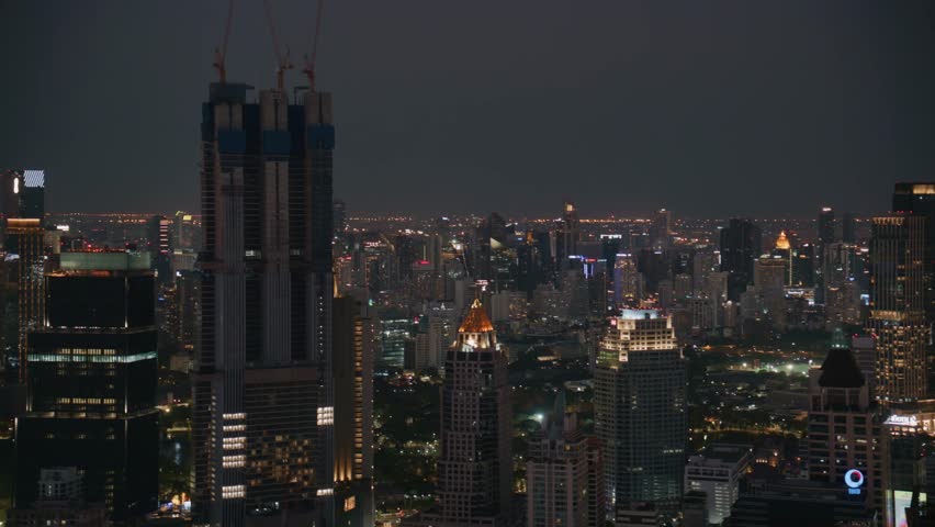Handheld Bangkok skyline at night with skyscraper under construction and lightning illuminating the sky over the city metropolis