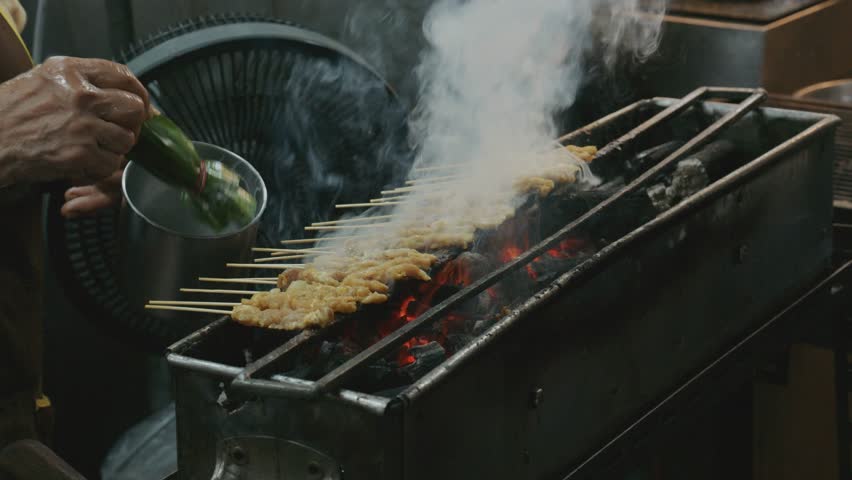 Street food vendor grills traditional pork satay skewers on a charcoal stove at a night market in Bangkok, Thailand
